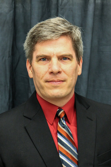 Image of Ole J. Forsberg. White male, grey hair. Dressed in a neat black suite and tie. 