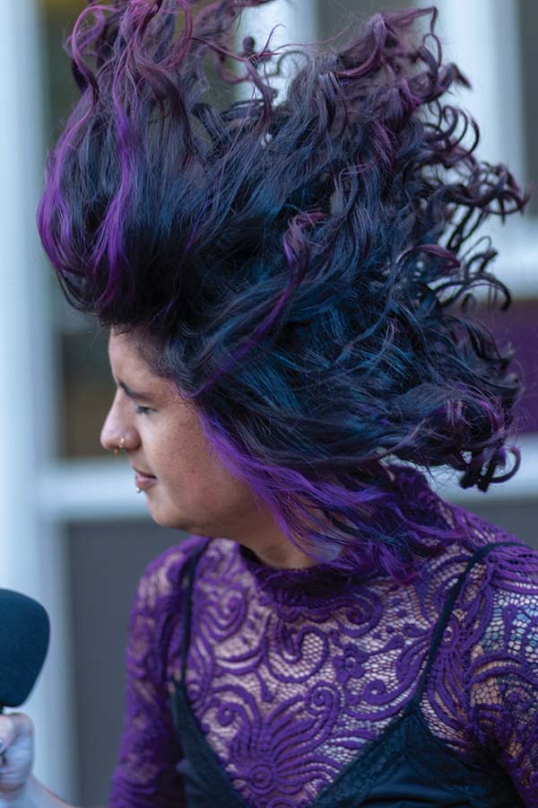 A singer wearing a purple shirt and with purple hair is flipping their hair around.