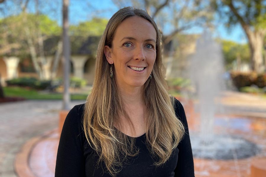 Lindsey stands in front of a water fountain with trees in the background wearing a black blouse, smiling at the camera.