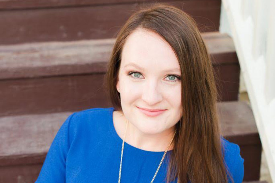 Erin who has long brown, straight hair wears a blue blouse and necklace and poses in front of a brown staircase.