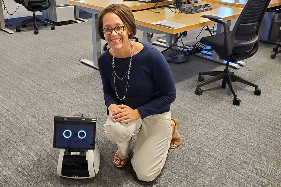 Rosie wears a blue blouse, brown glasses with a decorative, long necklace and light brown pants while kneeling next to Astro, a small robot with blue eyes lit on a screen. 