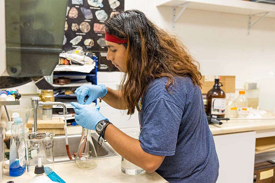 Juan stands over a beaker with a funnel connected to it near a sink in a science lab wearing a dark blue t-shirt, a red hair band, and blue latex gloves.