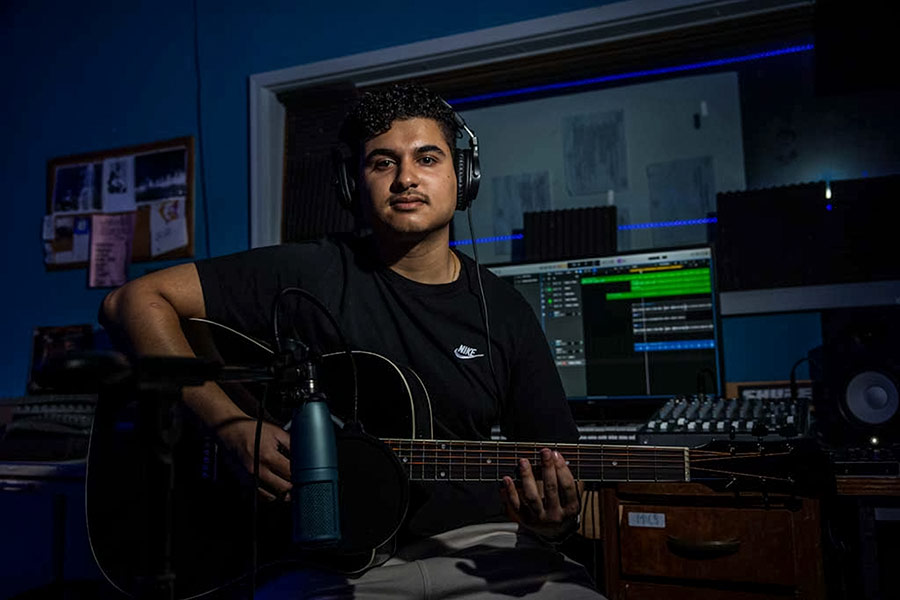 Neerav, in a black t-shirt and wearing black headphones, sits in front of DJ soundboard with a black, brown guitar next to a microphone.