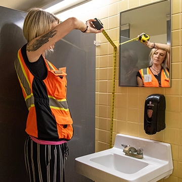 A student measures the height of a sink and mirror in a campus bathroom.