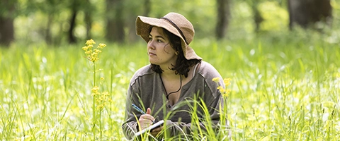 A student wearing a hat kneels in the middle of a field of prairie grass.