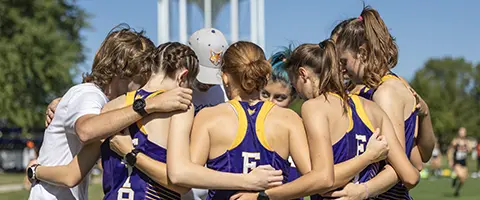 The Knox College women's cross country team huddles in a circle, arms around in each, in front of a water tower.