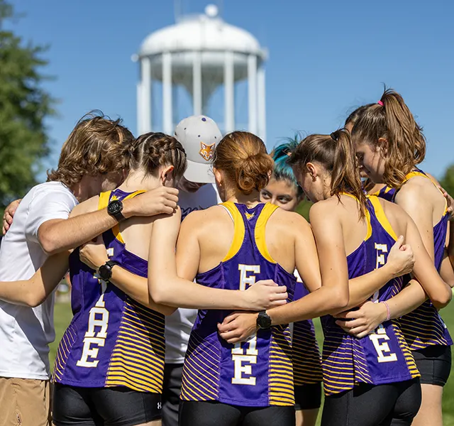 The Knox College women's cross country team huddles in a circle, arms around in each, in front of a water tower.