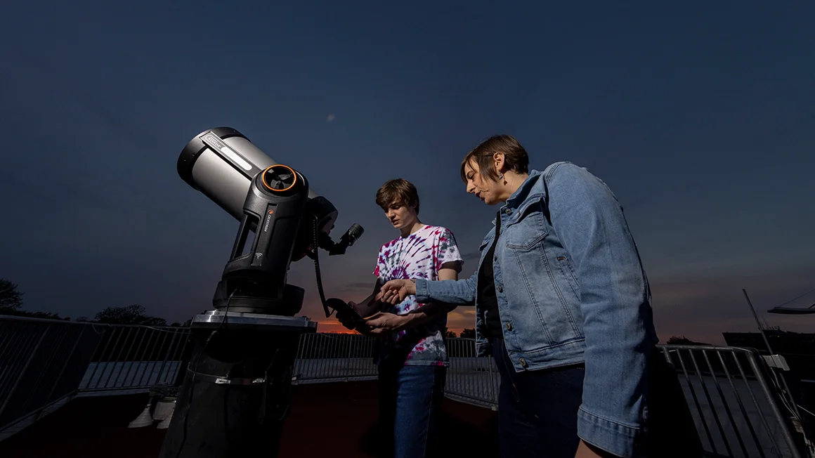 A Knox College faculty member and student stand at a telescope with the night sky behind and above them.