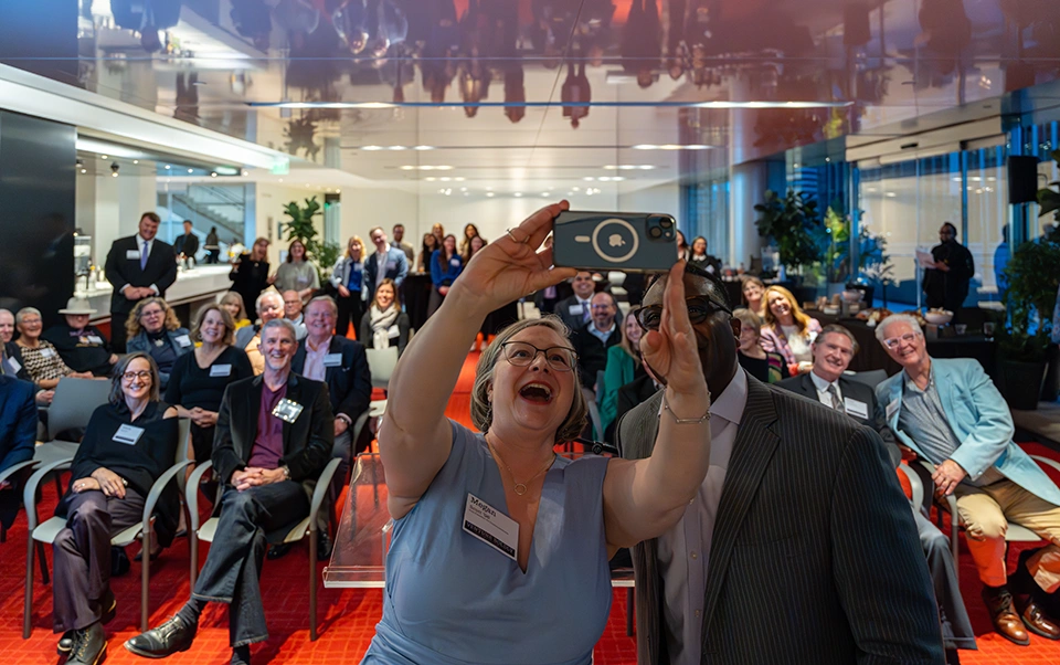 Megan Scott, Vice President for Communications, and President C. Andrew McGadney stand at the front of a room of people taking a selfie; event attendees smile for the photo in the background.