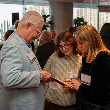 An event attendee in a light blue suit shows a book to two other event attendees who smile as they look at the book.