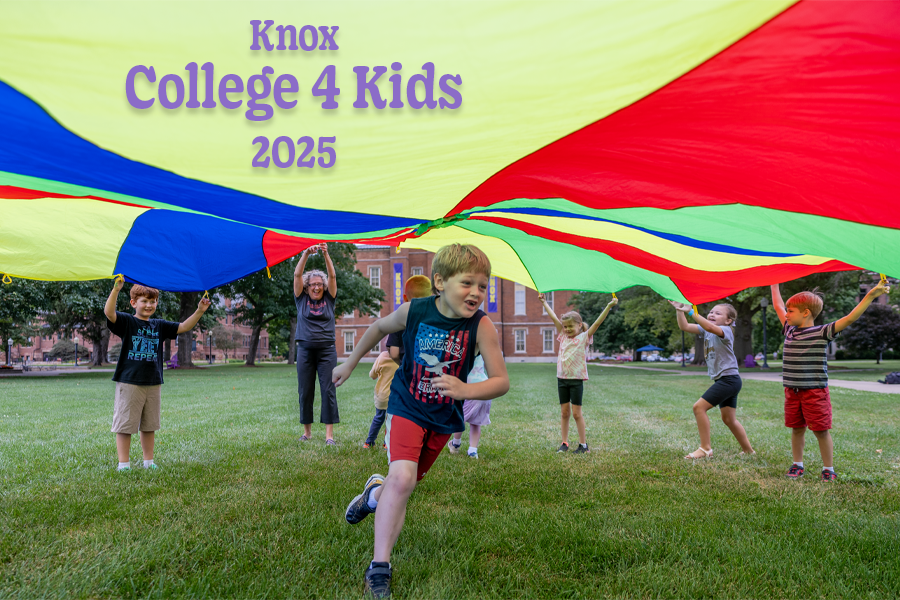 A child runs under a colorful parachute while other children and one adult hold the parachute over his head. Old Main is visible in the background.